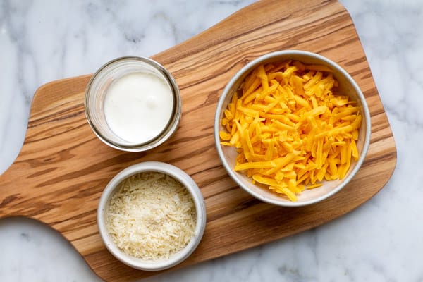 a bowl of shredded cheddar cheese, shredded parmesan cheese and heavy cream on a wooden board