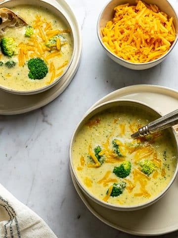 two bowls of broccoli soup; a bowl of cheese; a hand dipping bread