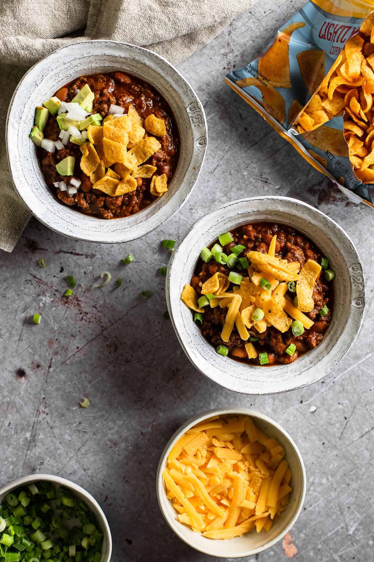 two bowls of chili, a bowl of cheese and an open bag of fritos