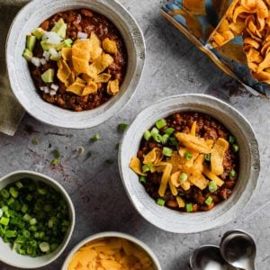 two bowls of chili topped with frito chips with green onion, cheese, and bag of fritos in background