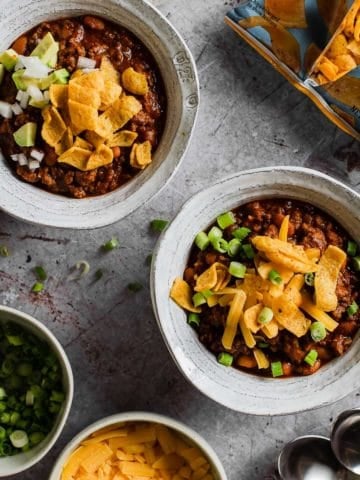 two bowls of chili topped with frito chips with green onion, cheese, and bag of fritos in background