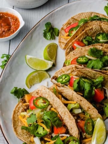 several tacos on a white platter with a bowl of salsa in the background