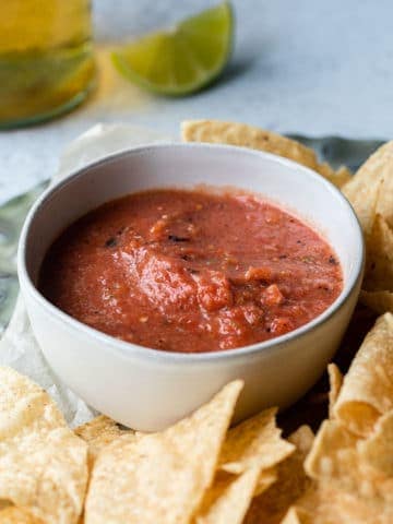 close up bowl of red salsa on a plate of chips with beer in background