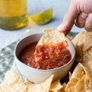 close up of hand dipping tortilla chip into a bowl of salsa with chips, beer. lime in background