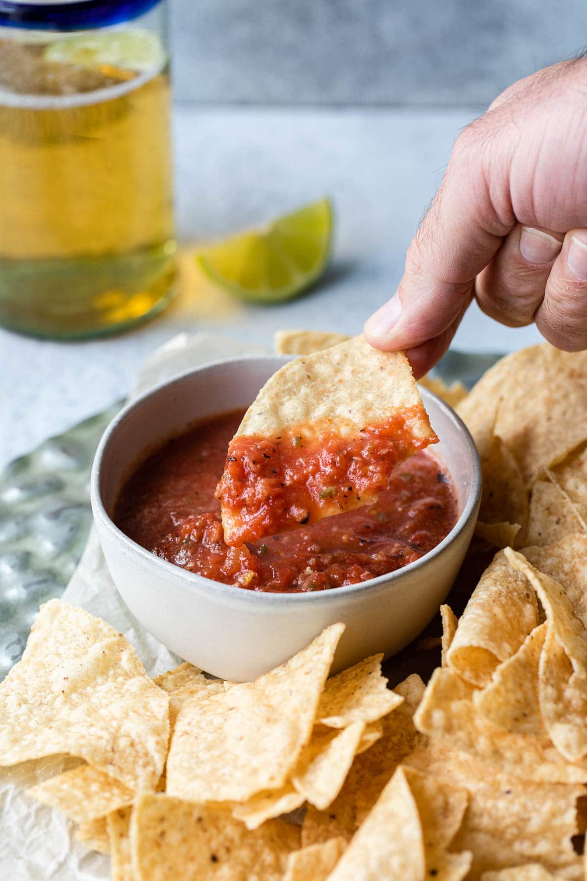 close up of hand dipping tortilla chip into a bowl of salsa with chips, beer. lime in background