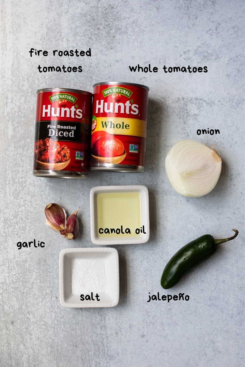 canned tomatoes, canola oil, jalepeno, garlic cloves, half and onion on a gray backgroudn