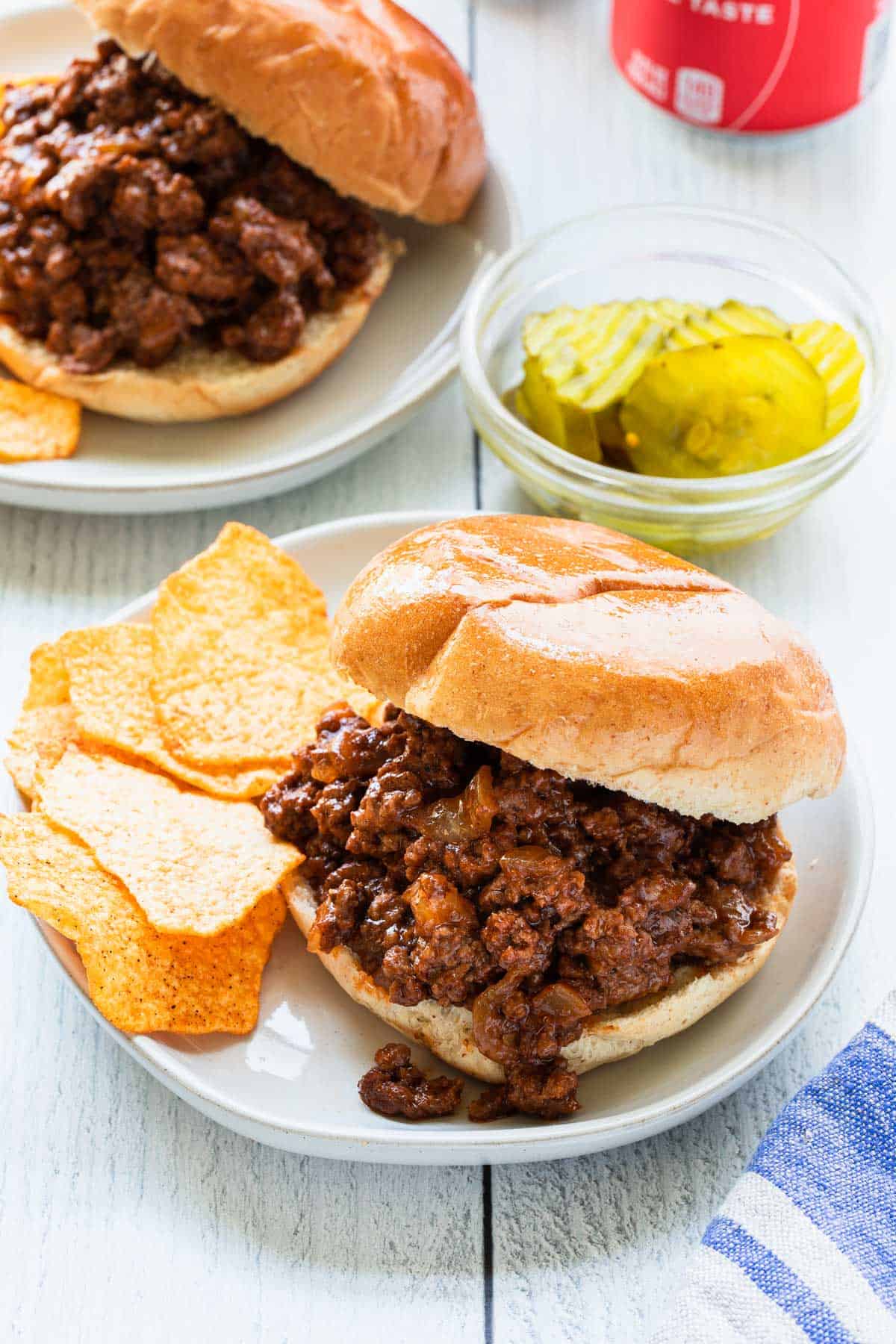 two plates of sloppy joes with a side of bbq chips, pickles