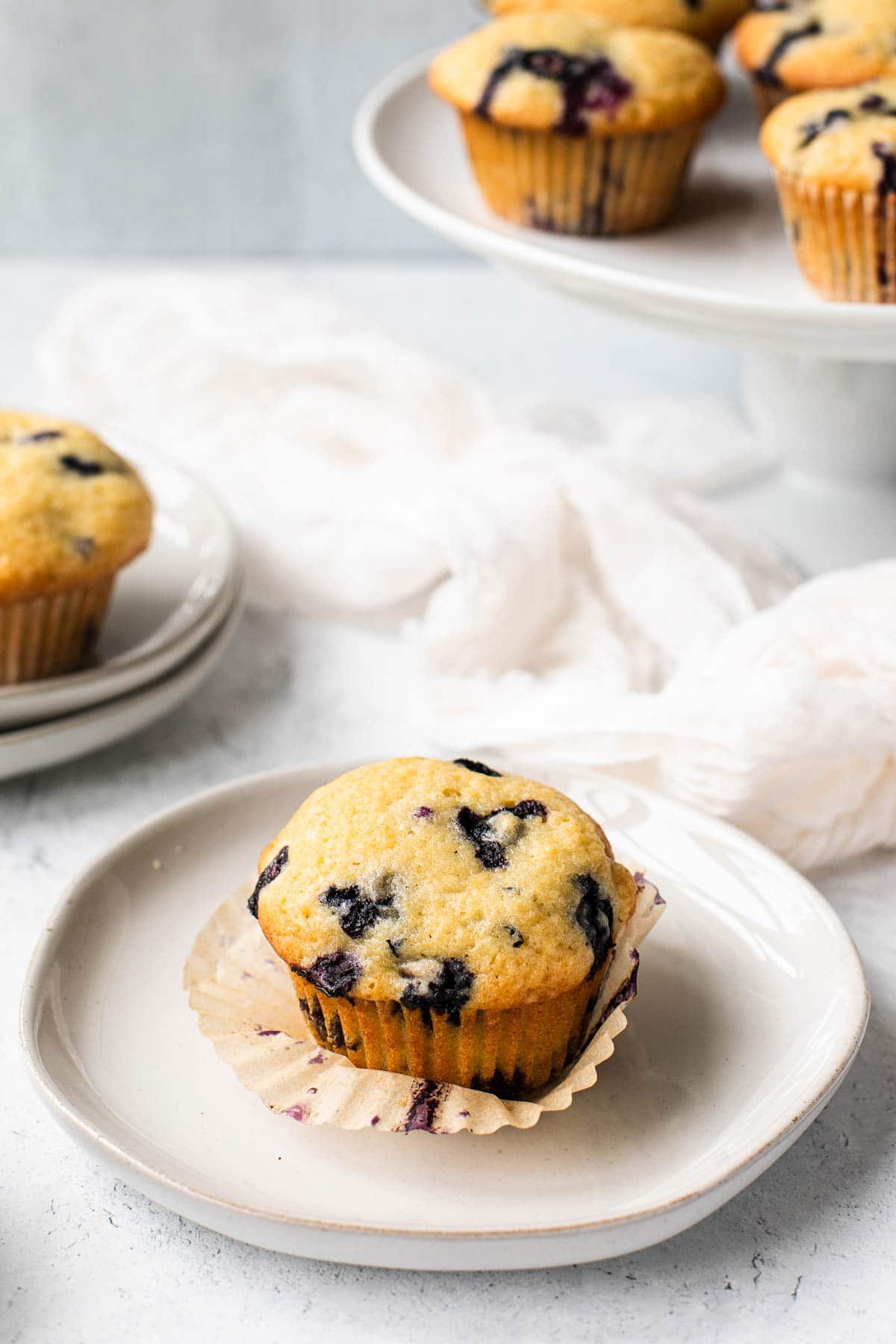 a blueberry muffin on a white plate with more muffins on a cake stand in the background