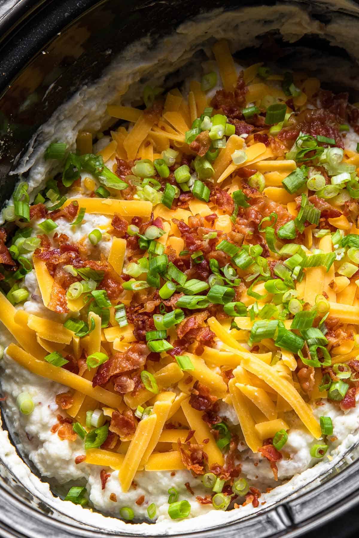 mashed potatoes in a crockpot loaded with shredded cheese, chopped bacon and green onions