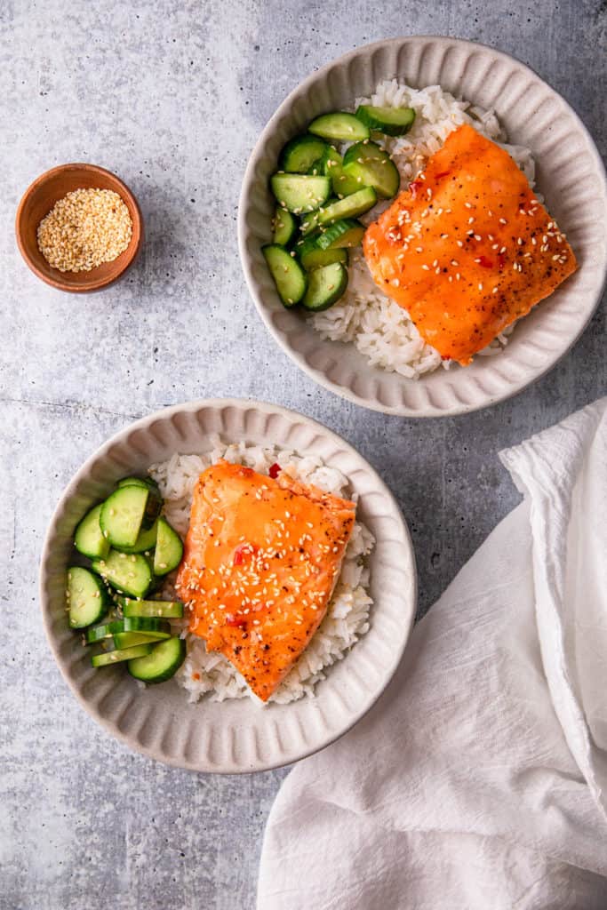 two bowls of sweet chili salmon over rice with cucumbers.