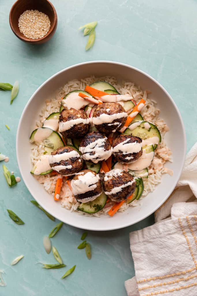 Abowl of meatballs over rice and cucumbers carrot salad. Creamy pink sauce drizzled over the top.