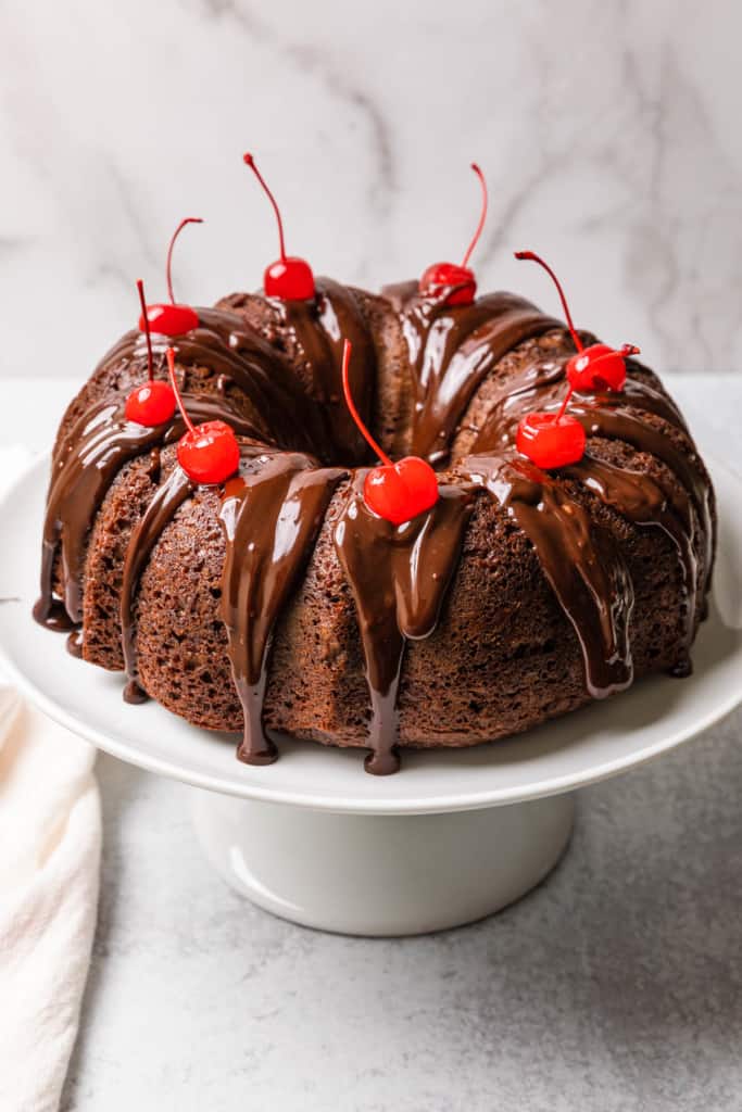 a chocolate bundt cake topped with cherries on a cake stand