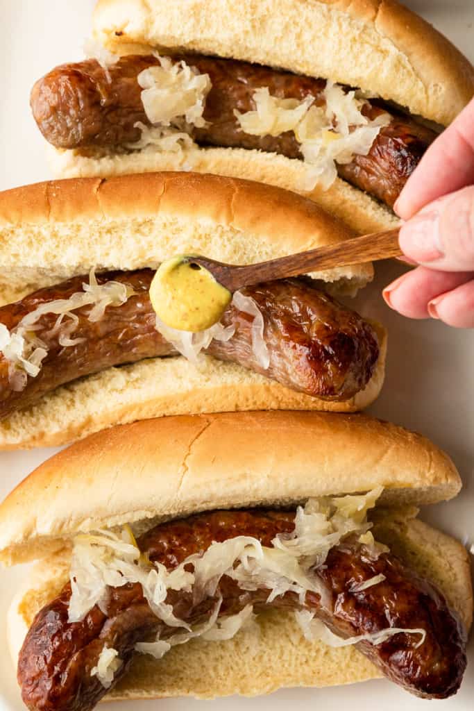 adding mustard to some bratwurst in buns with sauerkraut