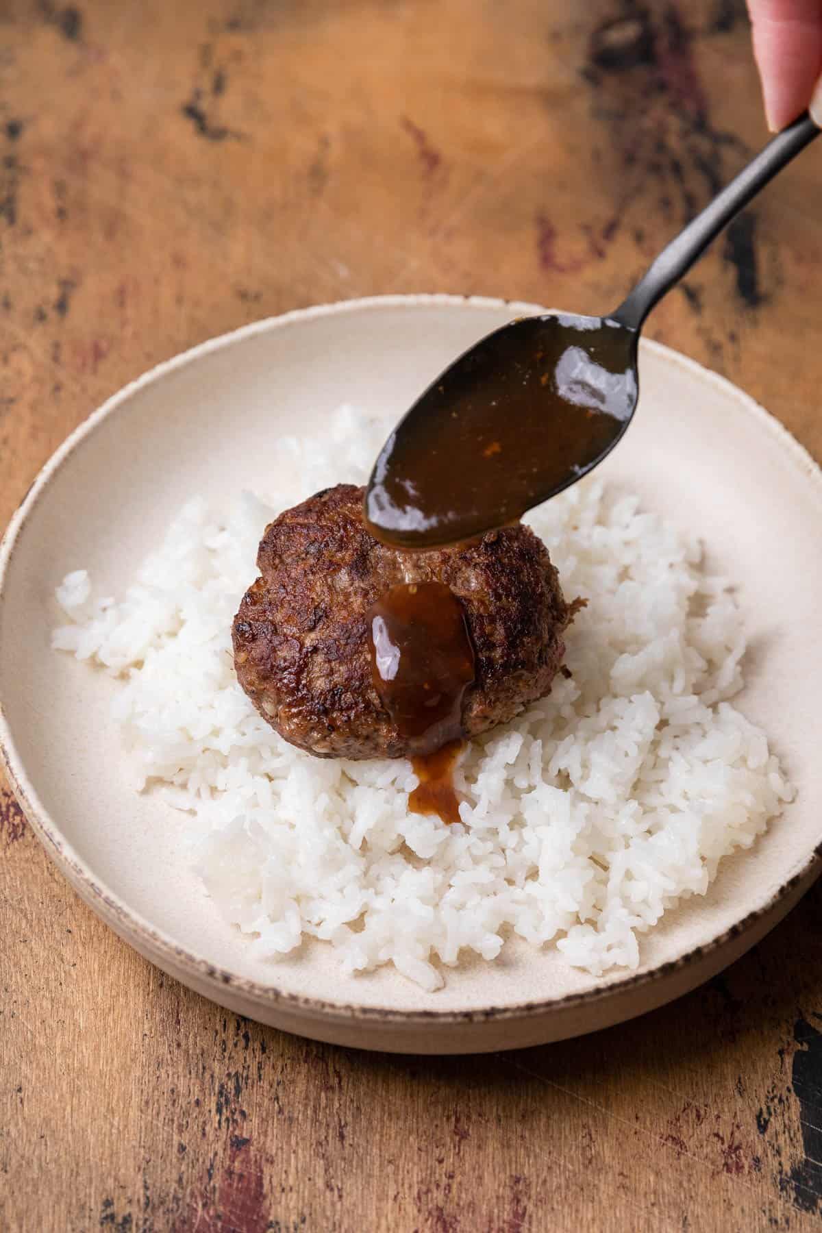 pouring gravy over a cooked hamburger patty over a bed of rice