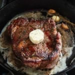 A ribeye cooking a cast iron skillet with a pat of herb and garlic butter on top.