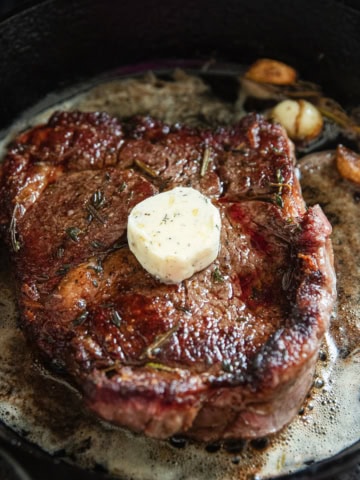 A ribeye cooking a cast iron skillet with a pat of herb and garlic butter on top.