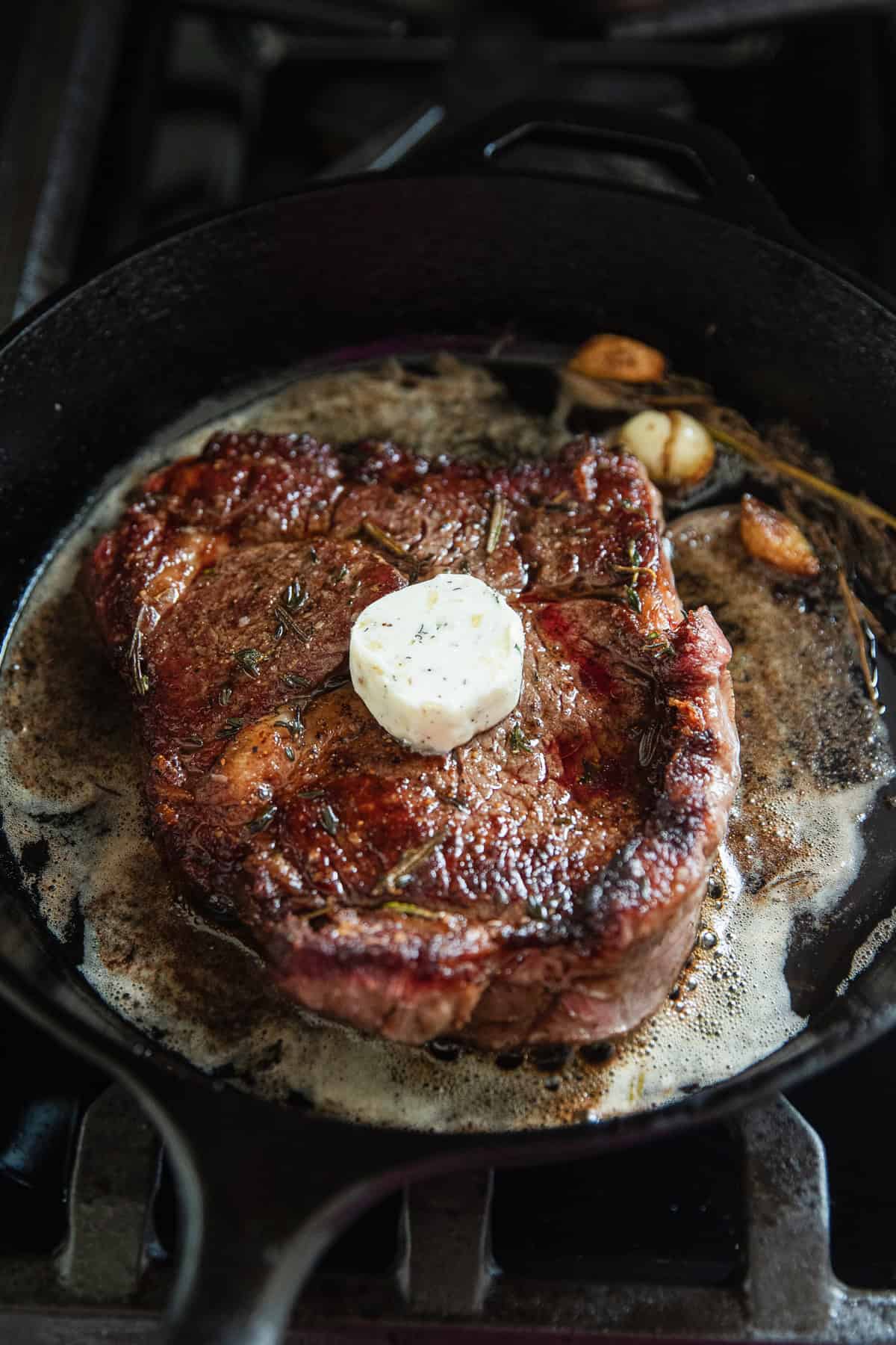 A ribeye steak cooking in a cast iron skillet with herb and garlic butter on top of steak.